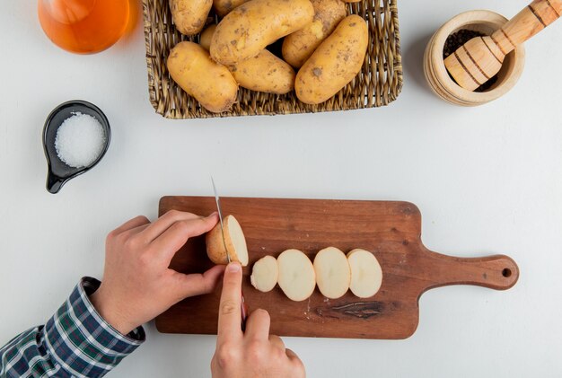 Vista superior de las manos cortando papas con un cuchillo en la tabla de cortar y otras en un plato de mantequilla, sal, pimienta negra sobre blanco