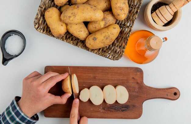 Vista superior de las manos cortando papas con un cuchillo en la tabla de cortar y otras en un plato de mantequilla, sal, pimienta negra sobre la superficie blanca