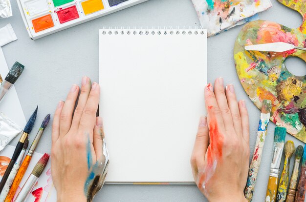 Vista superior manos con cuaderno rodeado por elementos de pintura