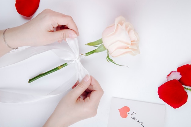 Vista superior de manos femeninas atando un lazo de cinta blanca en una flor rosa en el fondo blanco