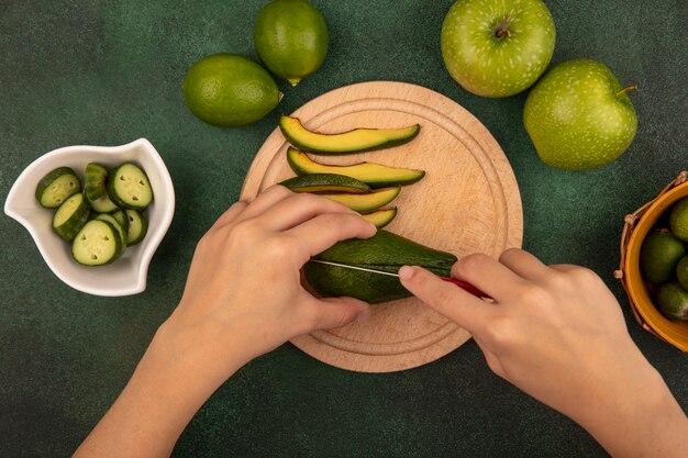 Vista superior de las manos femeninas cortando aguacate con un cuchillo en una tabla de cocina de madera con rodajas de pepino picado en un recipiente con limas y manzanas verdes aisladas sobre una superficie verde