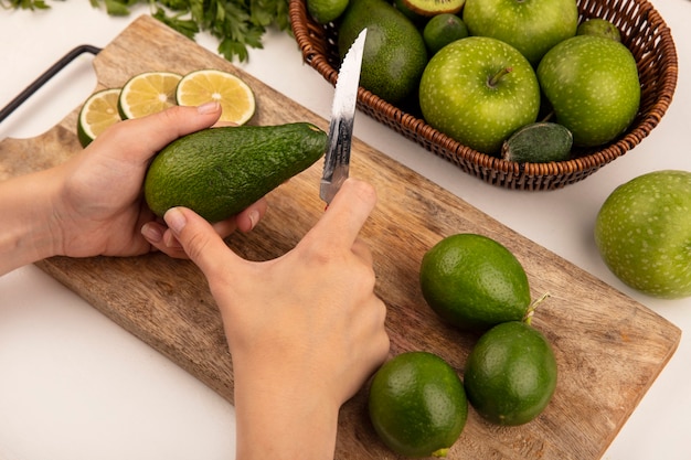 Vista superior de las manos femeninas cortando aguacate fresco con un cuchillo en una tabla de cocina de madera con limas con manzanas en un balde en una pared blanca