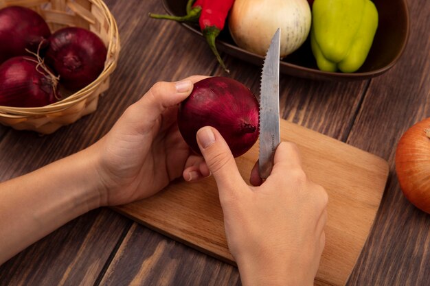 Vista superior de manos femeninas cortando una cebolla roja sobre una tabla de cocina de madera con un cuchillo en una pared de madera