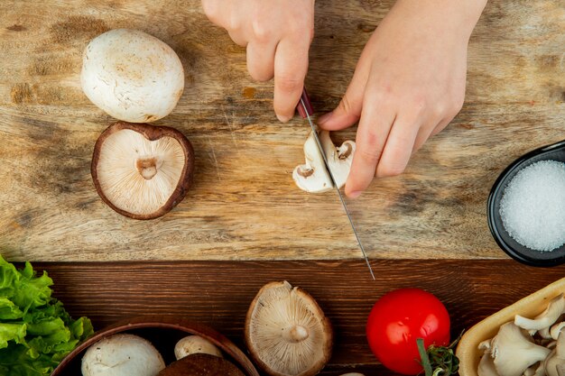 Vista superior de manos femeninas cortando champiñones frescos en tabla de cortar de madera y tomates frescos en mesa rústica de madera