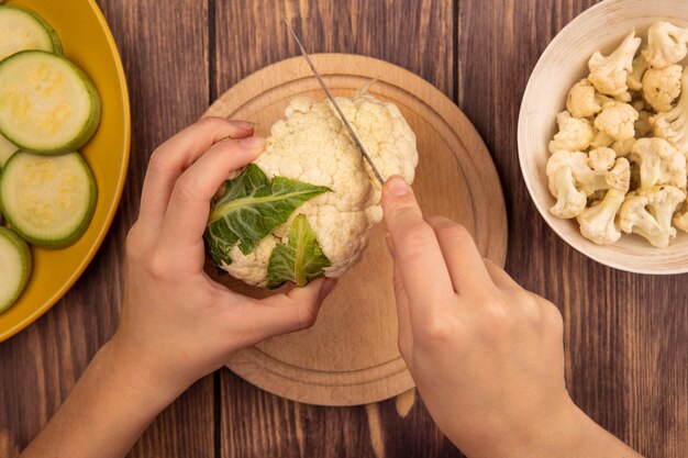 Vista superior de manos femeninas cortando coliflor en una tabla de cocina de madera con un cuchillo con cogollos de coliflor en un recipiente sobre una superficie de madera