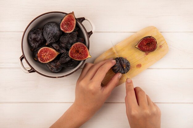 Vista superior de manos femeninas cortando higos negros dulces frescos en una tabla de cocina de madera con un cuchillo en una pared de madera blanca