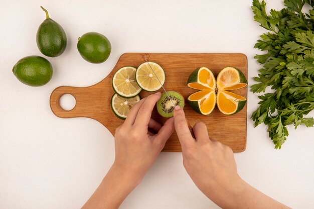 Vista superior de manos femeninas cortando kiwis frescos en una tabla de cocina de madera con cuchillo con feijoas aislado en una pared blanca