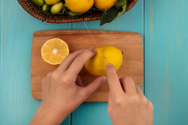 Vista superior de manos femeninas cortando limón fresco en una tabla de cocina de madera con un cuchillo con frutas como kinkans y limones en un cubo sobre una superficie de madera azul