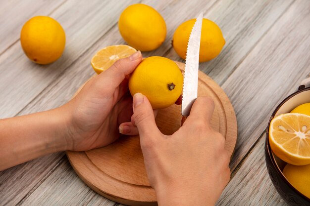 Vista superior de las manos femeninas cortando limón fresco en una tabla de cocina de madera con un cuchillo con limones en un recipiente con limones aislado sobre un fondo de madera gris