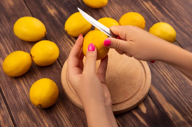 Vista superior de manos femeninas cortando limón fresco en una tabla de cocina de madera con cuchillo en una pared de madera