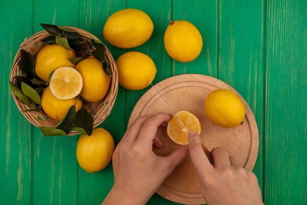 Vista superior de manos femeninas cortando limón en una tabla de cocina de madera con un cuchillo con limones en un balde en una pared de madera verde