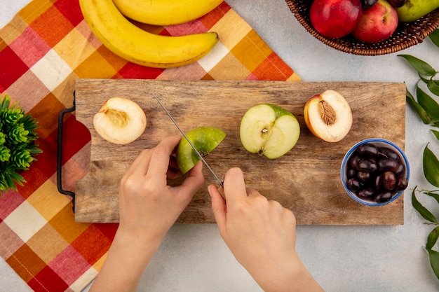 Vista superior de las manos femeninas cortando manzana con un cuchillo y medio corte melocotón y uva en la tabla de cortar y plátano en tela escocesa con melocotones y hojas sobre fondo blanco.