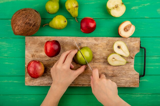 Vista superior de manos femeninas cortando manzana con cuchillo y medio corte pera y melocotones en tabla de cortar con coco sobre fondo verde