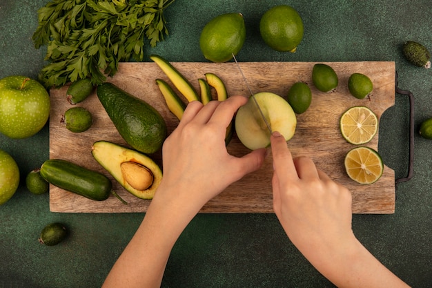 Vista superior de las manos femeninas cortando manzana fresca con un cuchillo en una tabla de cocina de madera con limas feijoas aguacates manzanas verdes y perejil aislado en una pared verde