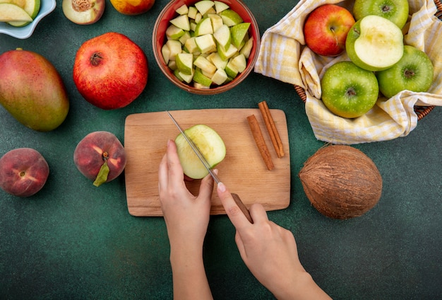 Vista superior de las manos femeninas cortando la manzana en rodajas en el tablero de la cocina con un cuchillo con palitos de canela, melocotón de coco aislado en verde