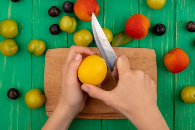 Vista superior de las manos femeninas cortando melocotón amarillo con un cuchillo sobre una tabla de cocina de madera sobre un fondo de madera verde