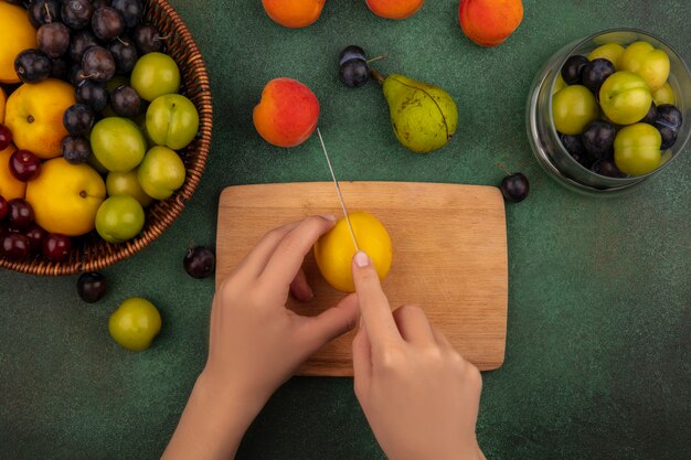 Vista superior de manos femeninas cortando melocotón amarillo con un cuchillo en una tabla de cocina de madera sobre un fondo verde