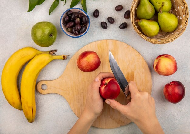 Vista superior de las manos femeninas cortando melocotón con un cuchillo en la tabla de cortar y manzana de plátano uva pera con hojas sobre fondo blanco.