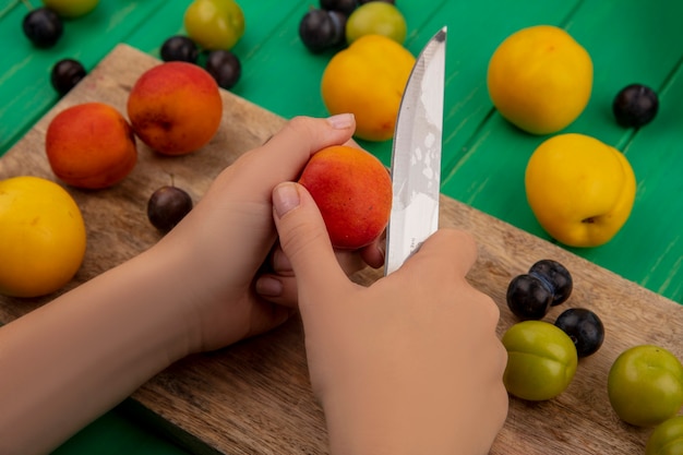 Vista superior de las manos femeninas cortando melocotón fresco con un cuchillo en una tabla de cocina de madera sobre un fondo verde