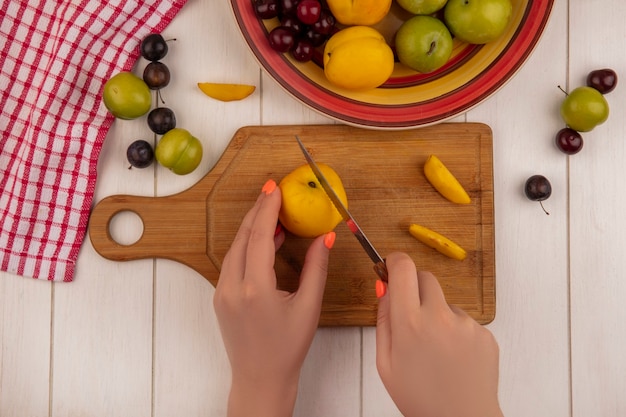 Foto gratuita vista superior de las manos femeninas cortando melocotón en un jabalí de cocina de madera con un cuchillo con frutas como ciruelas de cereza sloesgreen aisladas sobre un fondo blanco de madera