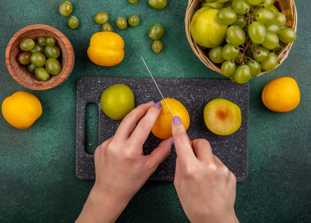 Vista superior de manos femeninas cortando nectacot con cuchillo y medio pluot cortado en tabla de cortar y pluot de uva en canasta con bayas de uva en un recipiente sobre fondo verde