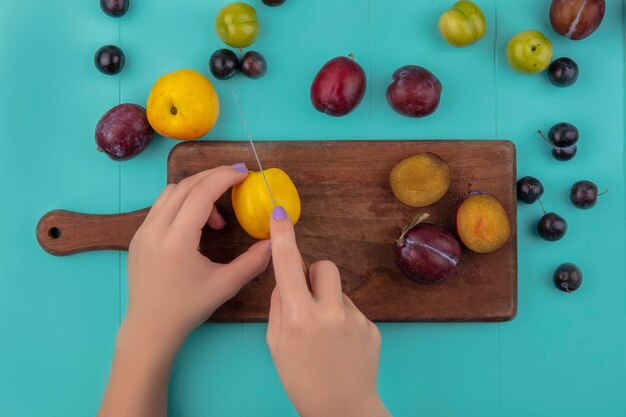 Vista superior de manos femeninas cortando nectacot con cuchillo y pluots sobre tabla de cortar y patrón de nectacot de uva plum pluot sobre fondo azul.