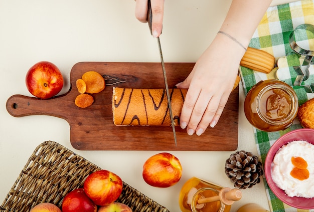 Vista superior de manos femeninas cortando el pastel sobre una tabla de cortar de madera y frutas frescas nectarinas y duraznos con un vaso de mermelada de durazno y queso