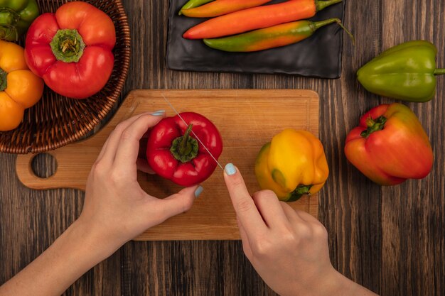 Vista superior de manos femeninas cortando pimiento aromático fresco en una tabla de cocina de madera con un cuchillo sobre una superficie de madera