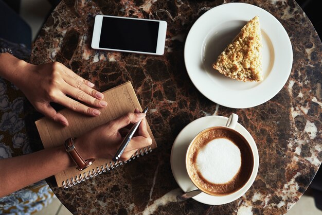 Vista superior de manos femeninas haciendo notas en un café y postre