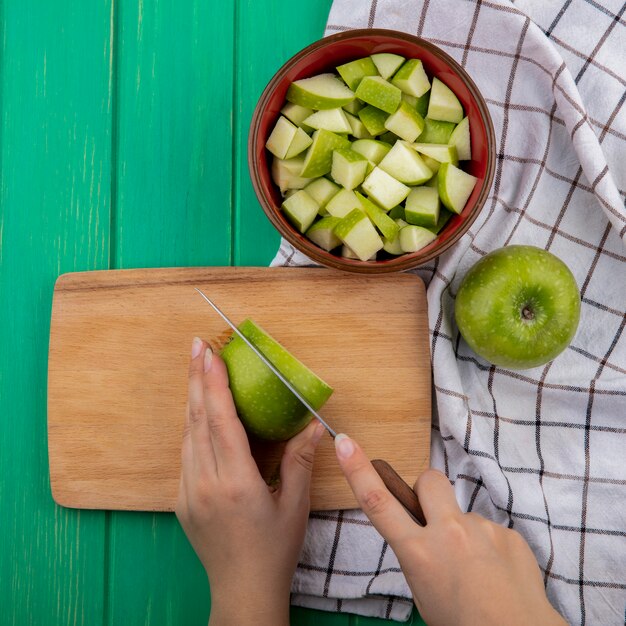 Vista superior de las manos femeninas para picar manzanas verdes sobre una placa de cocina de madera en un tazón rojo de manzanas picadas y tela