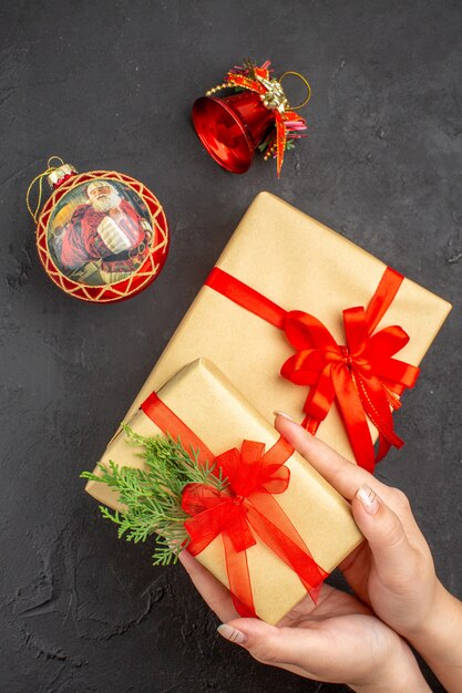 Vista superior de las manos femeninas con regalo de Navidad en papel marrón atado con cinta roja juguetes de árbol de Navidad en superficie oscura