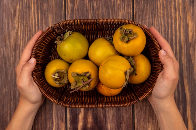 Vista superior de manos femeninas sosteniendo un balde de frutas frescas de caqui naranja en una mesa de madera