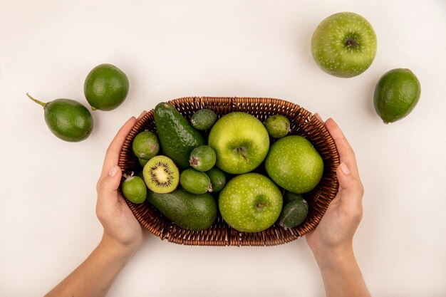 Vista superior de manos femeninas sosteniendo un balde de frutas frescas como manzanas, feijoas y aguacate sobre una superficie blanca