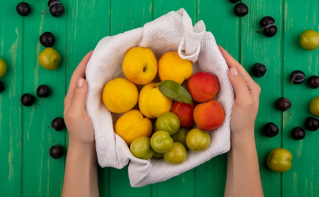 Foto gratuita vista superior de manos femeninas sosteniendo un balde con frutas frescas como melocotones ciruelas cereza verde sobre un fondo verde