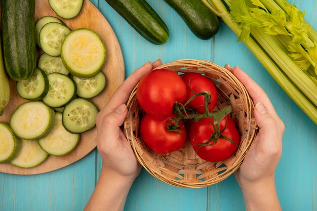 Foto gratuita vista superior de manos femeninas sosteniendo un balde de tomates frescos con pepinos picados y calabacines en una tabla de cocina de madera con apio aislado en una pared de madera azul