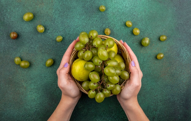 Vista superior de manos femeninas sosteniendo la cesta de frutas como uvas y pluots con uvas sobre fondo verde