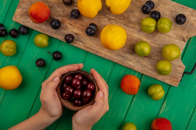 Vista superior de manos femeninas sosteniendo un cuenco de madera con cerezas rojas con melocotones amarillos con endrinas en una tabla de cocina de madera sobre un fondo verde