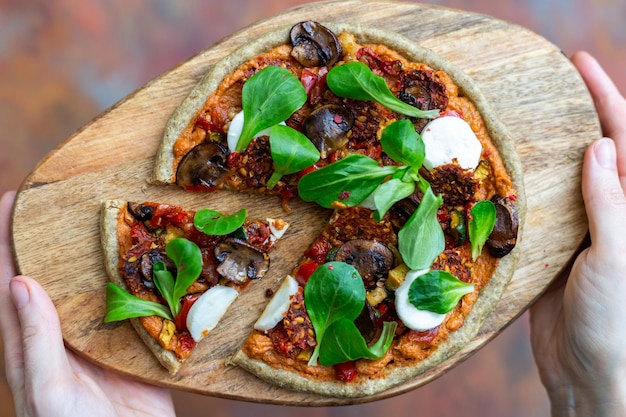 Vista superior de manos femeninas sosteniendo una pizza de champiñones vegana cruda servida en una tabla de madera