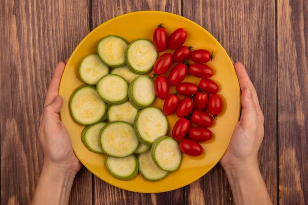 Vista superior de manos femeninas sosteniendo un plato amarillo de verduras frescas como tomates y calabacines sobre una superficie de madera