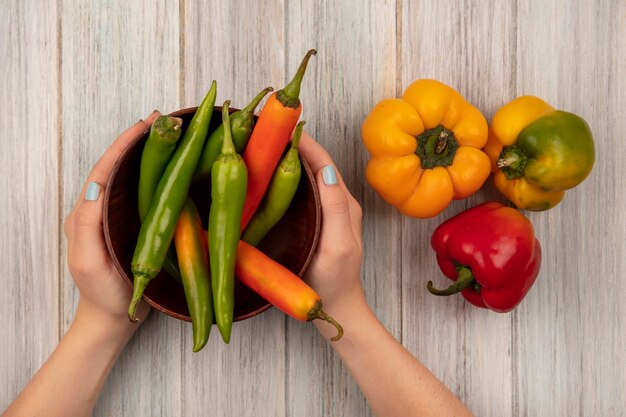 Vista superior de manos femeninas sosteniendo un tazón de pimientos con pimientos aislados sobre una superficie de madera gris