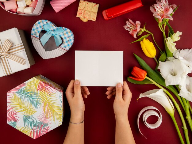 Vista superior de manos femeninas con tarjeta de felicitación de papel en blanco sobre mesa roja con tulipanes de color rojo y amarillo con alstroemeria y caja de regalo en forma de corazón y chocolate blanco