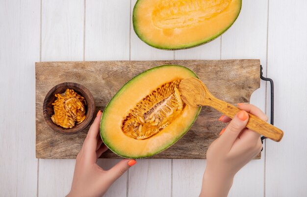 Vista superior de manos femeninas tomando semillas de melón con una cuchara de madera sobre tablero de cocina de madera con melón cantalupo en madera blanca