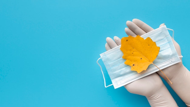 Vista superior de manos con guantes sosteniendo mascarilla médica con hoja de otoño