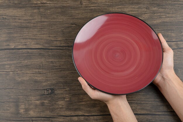 Vista superior de las manos del hombre sosteniendo un plato rojo vacío sobre una mesa de madera.