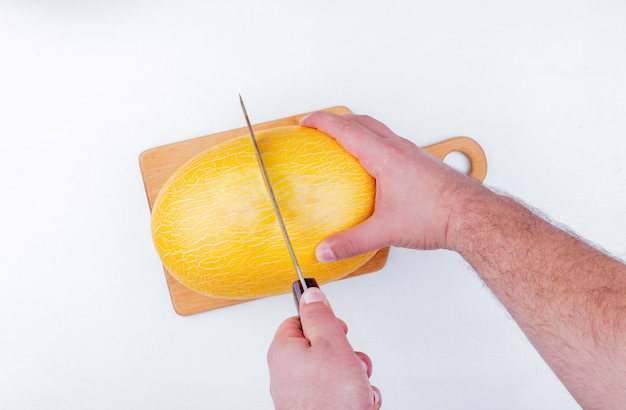 Vista superior de manos masculinas cortando melón con cuchillo en la tabla de cortar sobre fondo blanco.