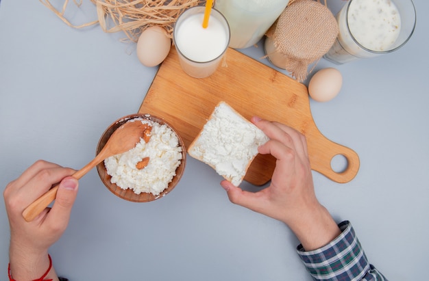 Vista superior de manos masculinas que sostienen una rebanada de pan untada con requesón y una cuchara con leche en la tabla de cortar y huevos, yogur, sopa, crema, paja en la mesa azul