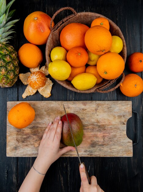 Vista superior de manos de mujer cortando mango con cuchillo en la tabla de cortar y frutas cítricas como naranja, limón, mandarina, piña en la mesa de madera