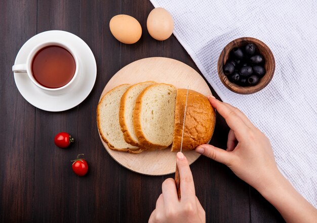 Vista superior de las manos de mujer cortando pan con un cuchillo en la tabla de cortar y una taza de té huevos tomates con tazón de aceituna negra sobre tela sobre superficie de madera