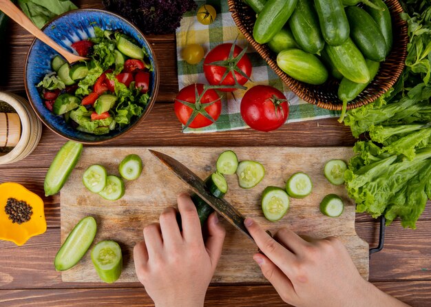 Vista superior de las manos de mujer cortando pepino con un cuchillo en la tabla de cortar con ensalada de lechuga, tomate, pimienta negra sobre superficie de madera