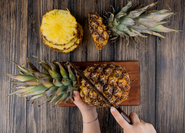 Vista superior de manos de mujer cortando piña con un cuchillo en la tabla de cortar con rodajas de piña en la mesa de madera